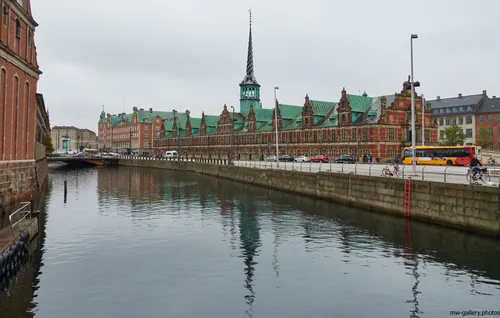 This urban landscape photograph captures the architecture of Copenhagen with a view of the old stock exchange canal during a late morning in autumn. The main subject is the historic Børsen building, identifiable by its distinctive green copper roof and spiral spire, prominently positioned alongside the canal. The city’s architectural heritage is evident in the traditional style of the buildings lining the waterfront, while modern elements such as parked cars, bicycles, and a public bus highlight the contemporary urban life of Copenhagen. The overcast sky and subdued light are characteristic of the autumn season, and the canal’s reflection adds depth to the composition. This scene showcases the blend of old and new found in Copenhagen's city center, emphasizing its architectural significance within an active urban environment.